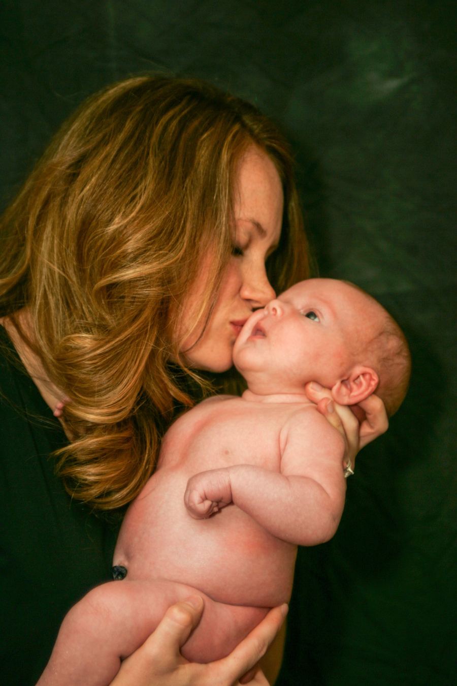A mother kisses her newborn baby against a deep green backdrop